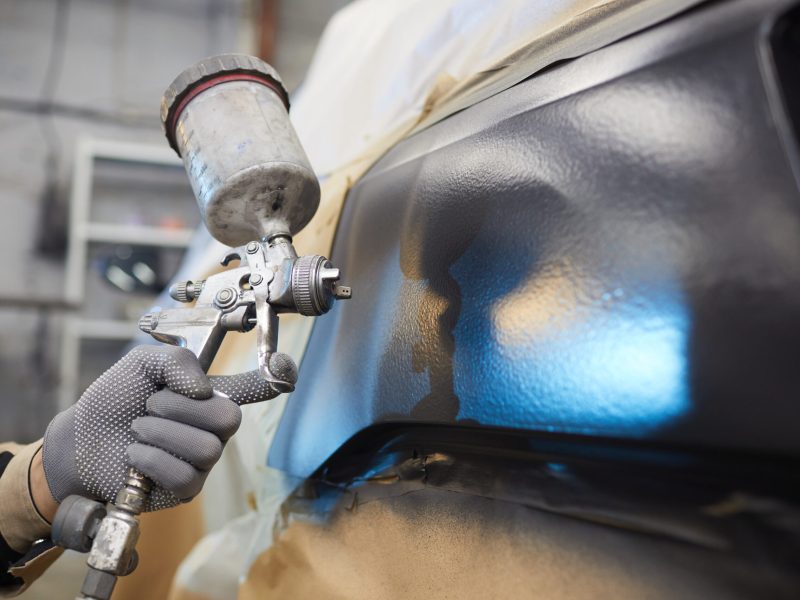 Male hand in protective glove holding spay-gun painting car body, horizontal close up shot, copy space